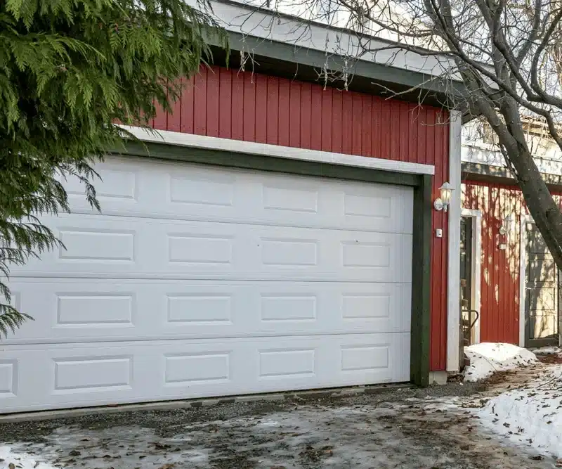 White garage door on gravel driveway with snow, red siding home in winter
