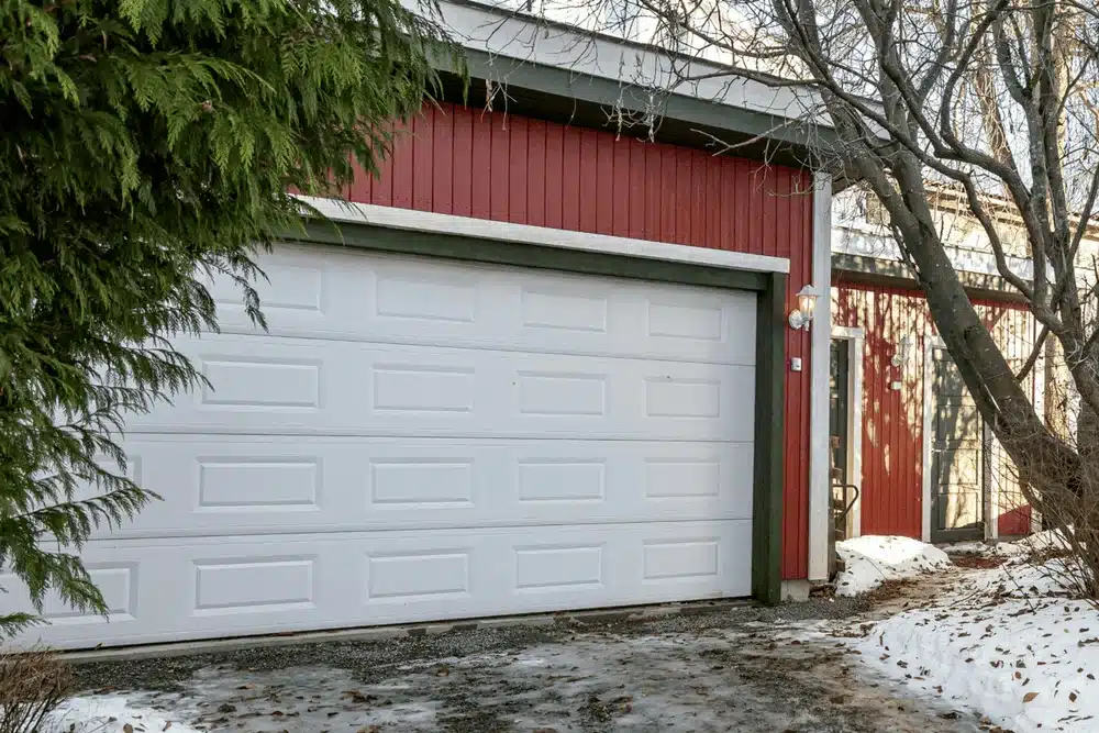 White garage door on gravel driveway with snow, red siding home in winter